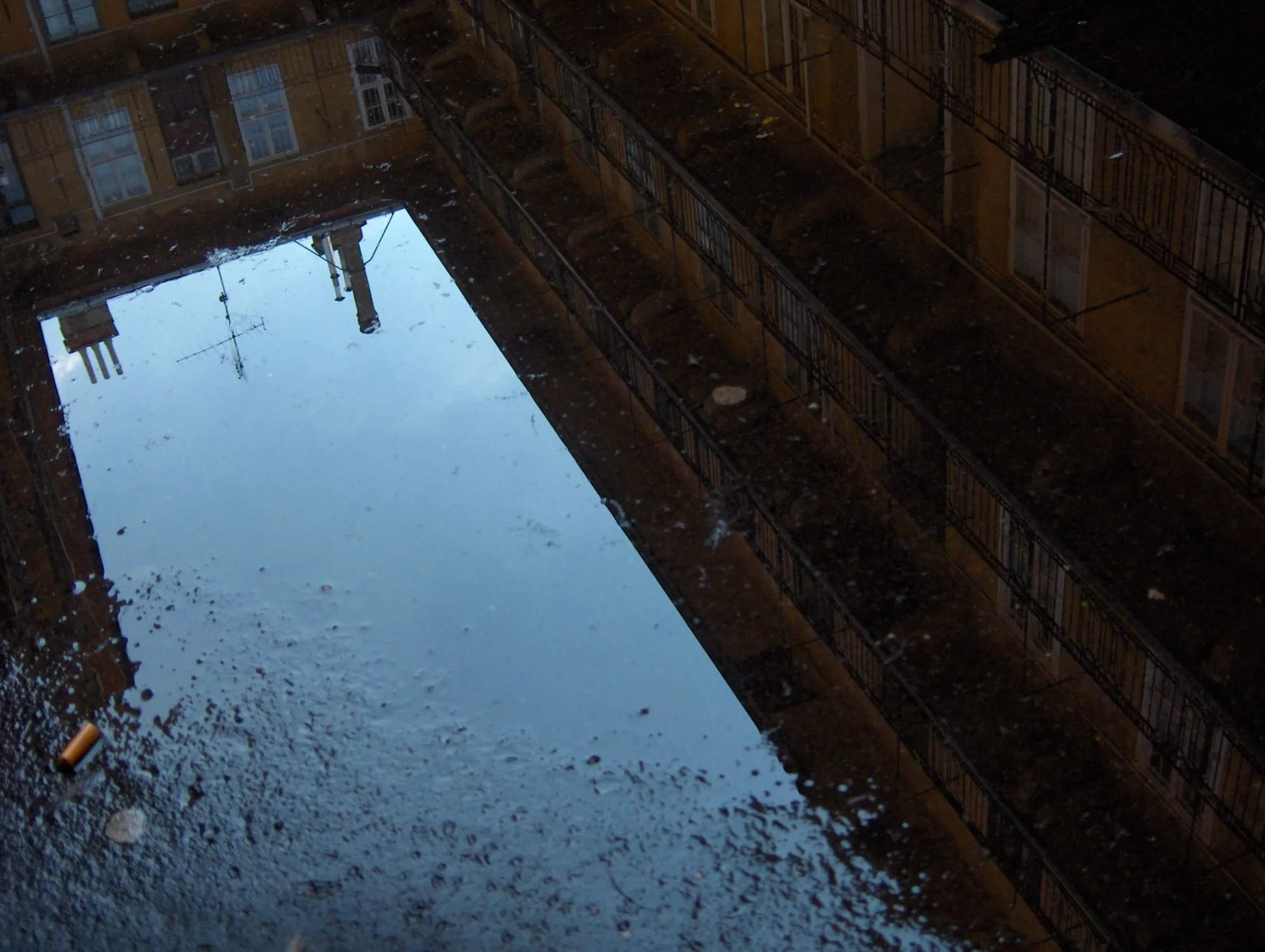 Patio de vecinos y cielo reflejados en un charco, con una colilla en la esquina