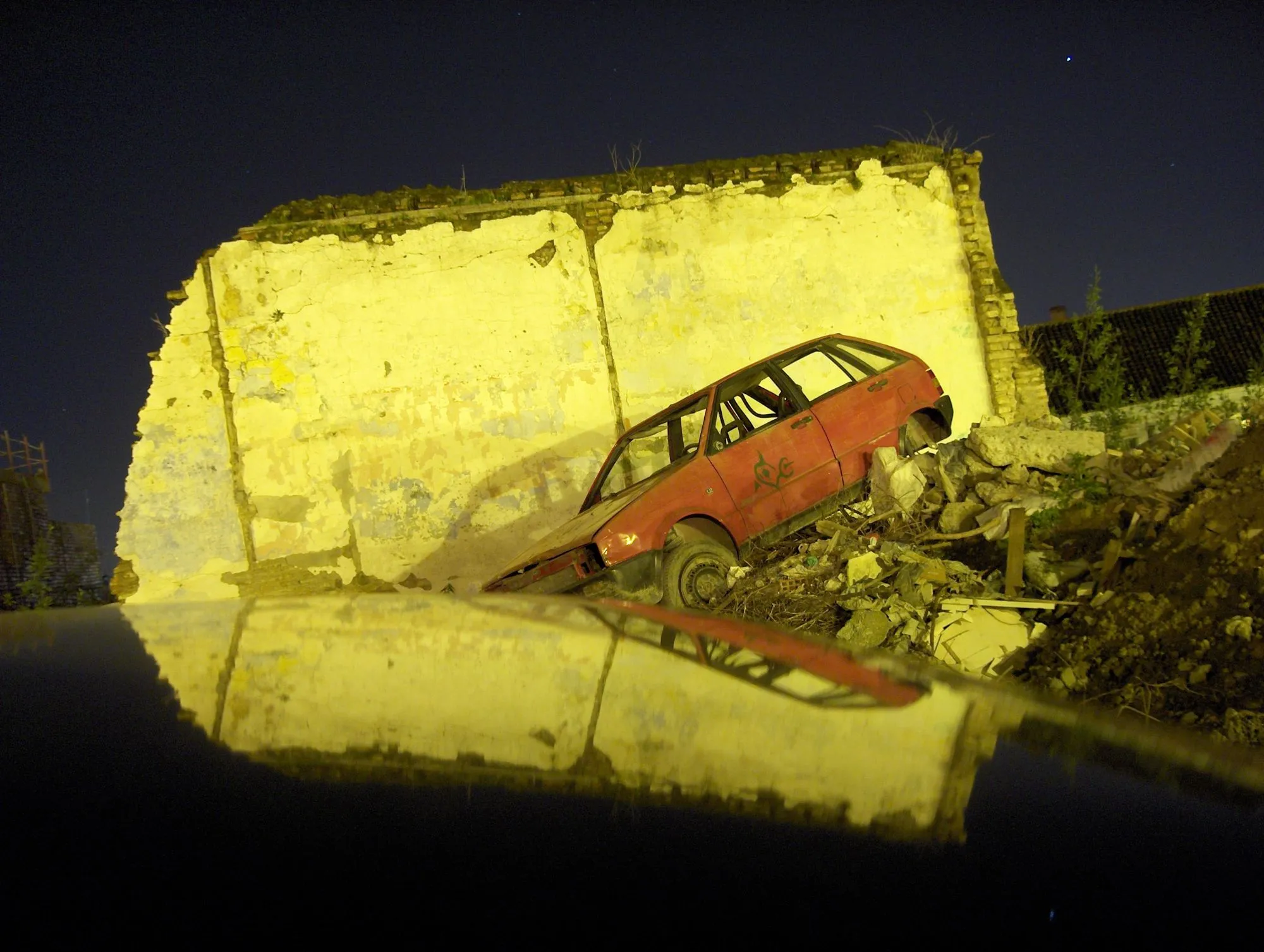 Coche rojo abandonado sobre escombros junto a un muro amarillo de noche, reflejado en un capó