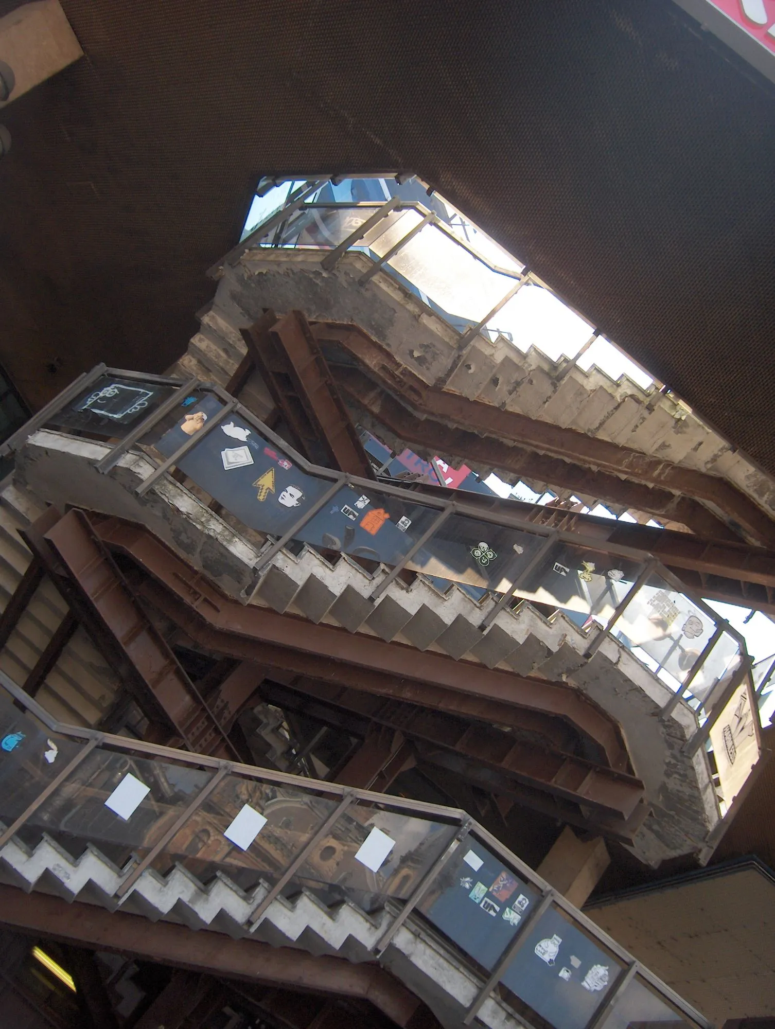 Escaleras de hormigón y vigas oxidadas vistas desde abajo, con pegatinas en las barandillas
