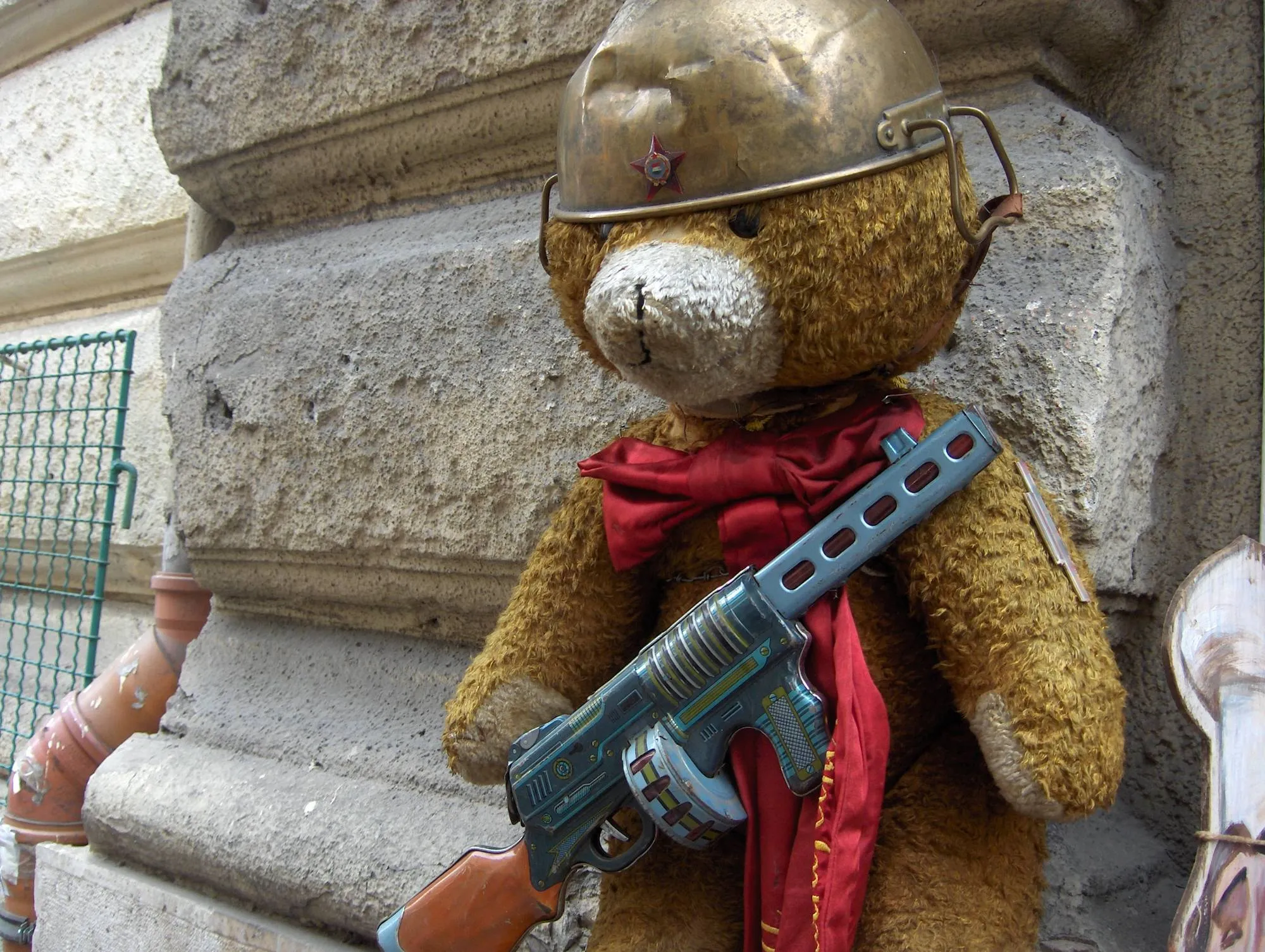 Oso de peluche con casco, pañuelo rojo y metralleta de juguete apoyado en una fachada de piedra de Budapest