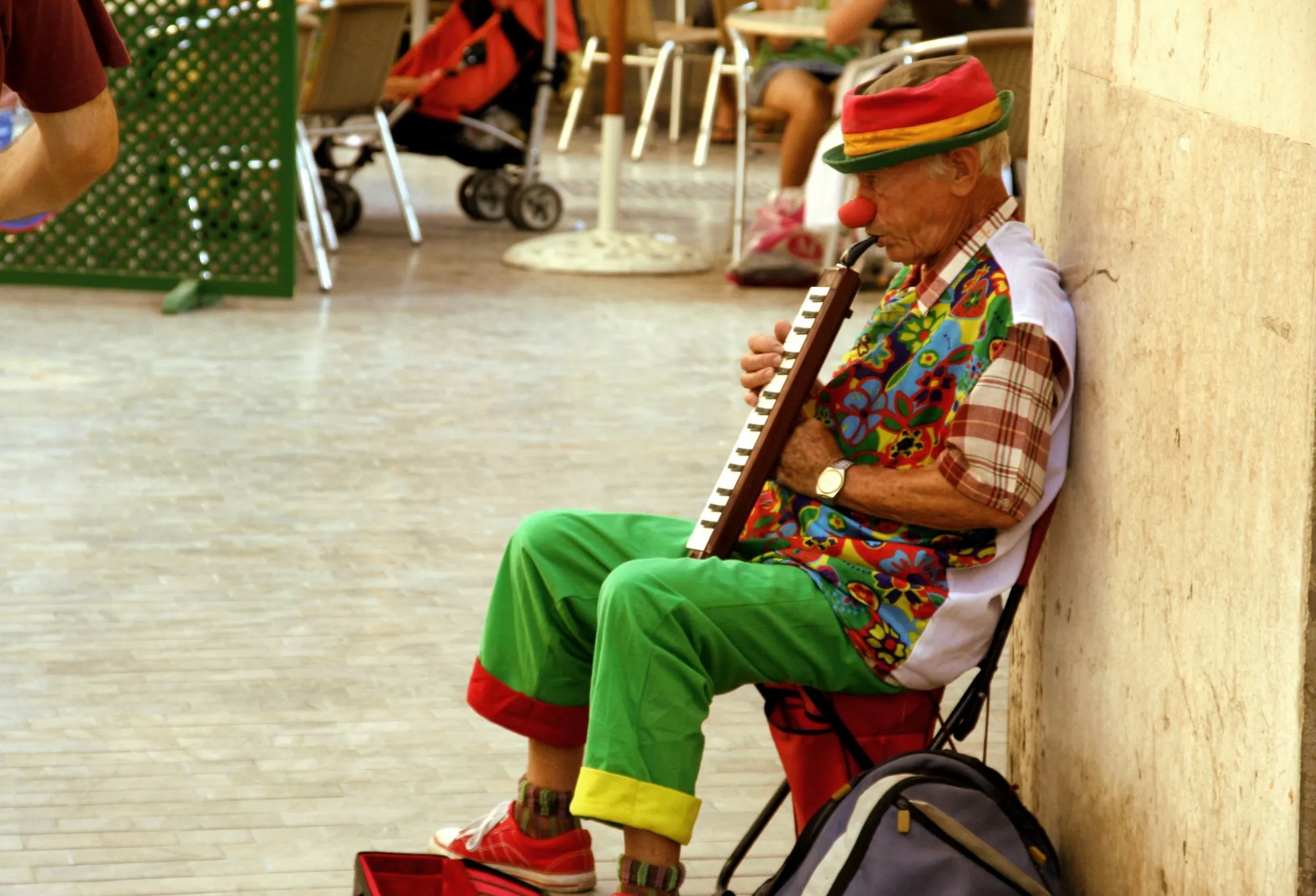 Payaso mayor con nariz roja tocando una melódica sentado junto a una terraza