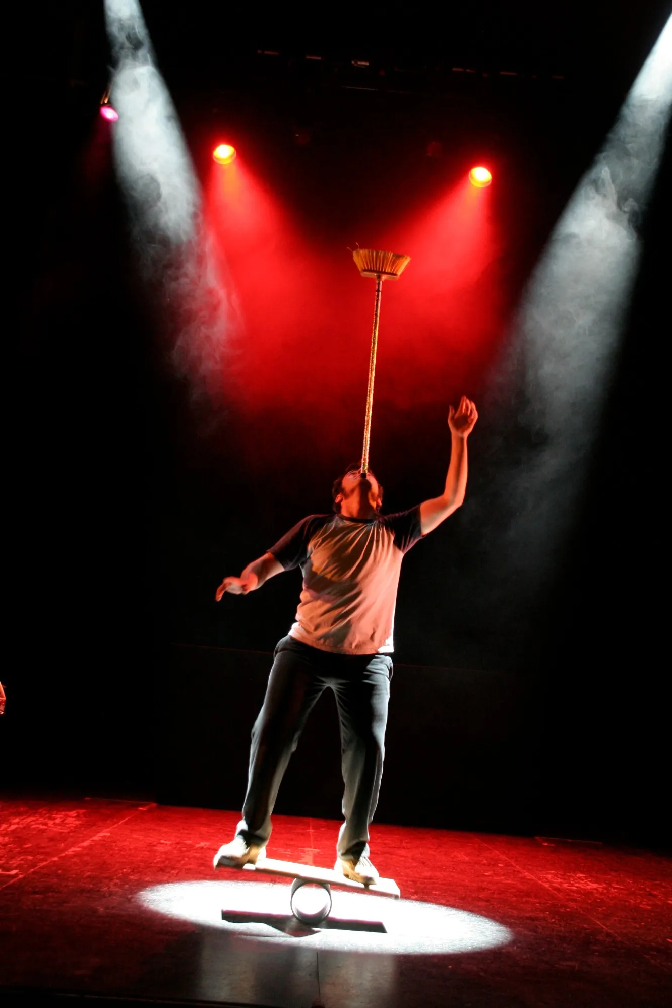 Equilibrista sobre una tabla en el escenario con una escoba en equilibrio sobre la barbilla, entre focos rojos y humo