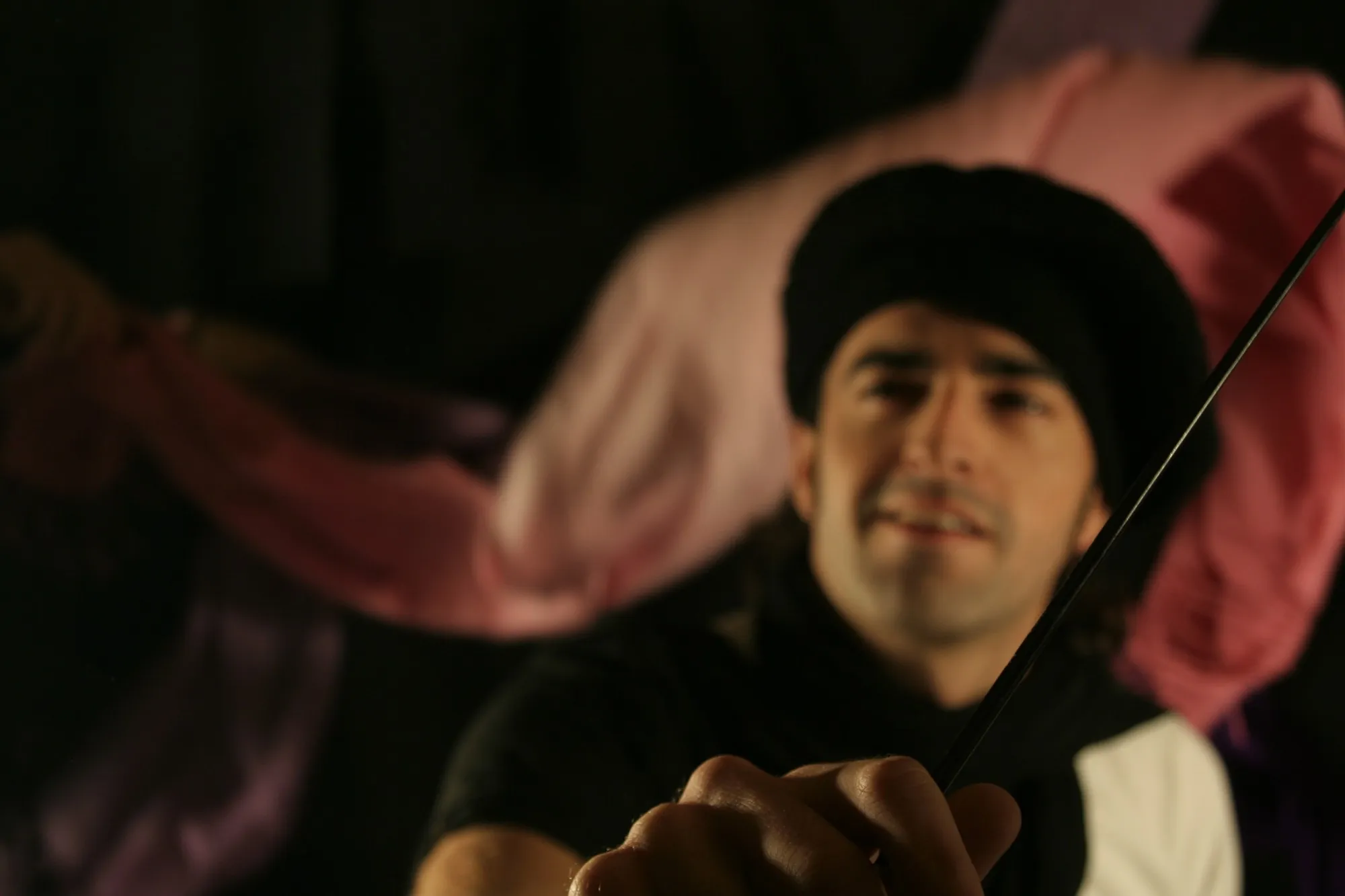 Hombre con gorro negro sujetando una varilla, desenfocado detrás de su propia mano, con telas rosas de fondo