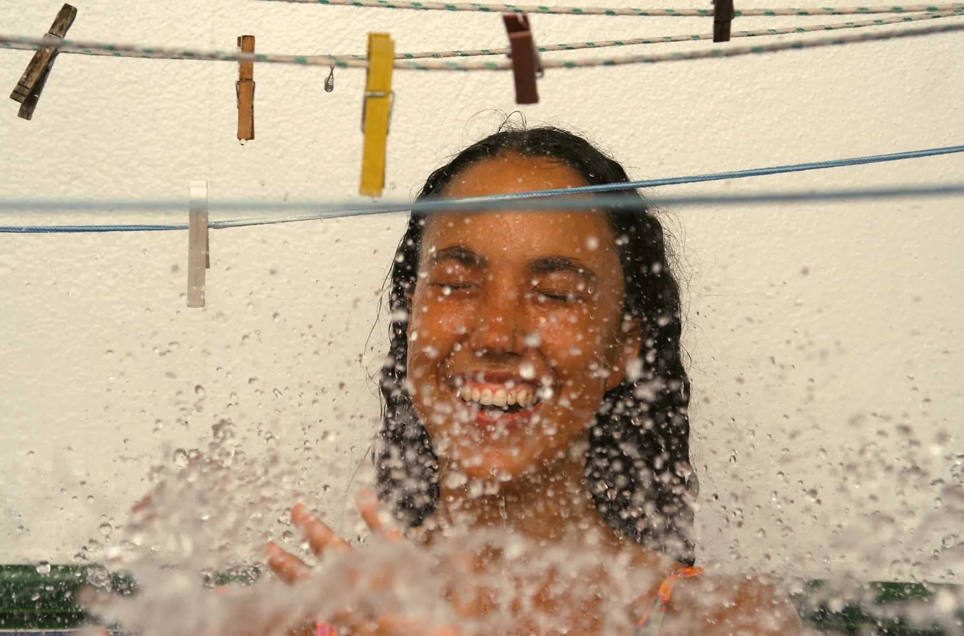 Chica riendo con los ojos cerrados bajo un chorro de agua, entre cuerdas de tender con pinzas