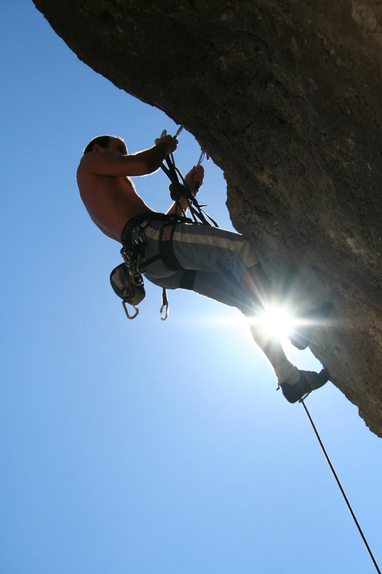 Escalador colgado de una roca desplomada a contraluz, con el sol entre las piernas