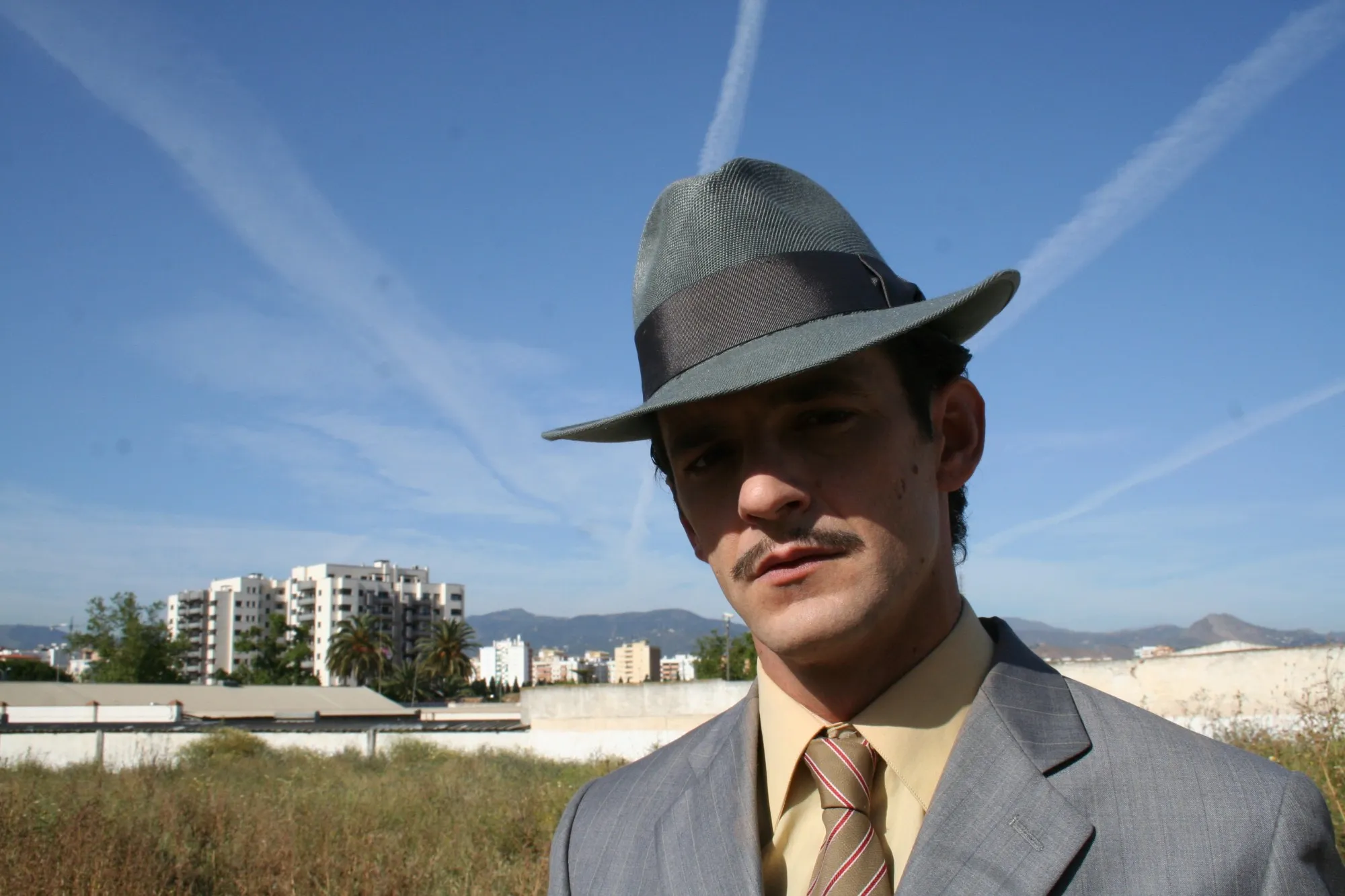 Hombre con sombrero gris, bigote y traje en un descampado de Málaga bajo estelas de avión
