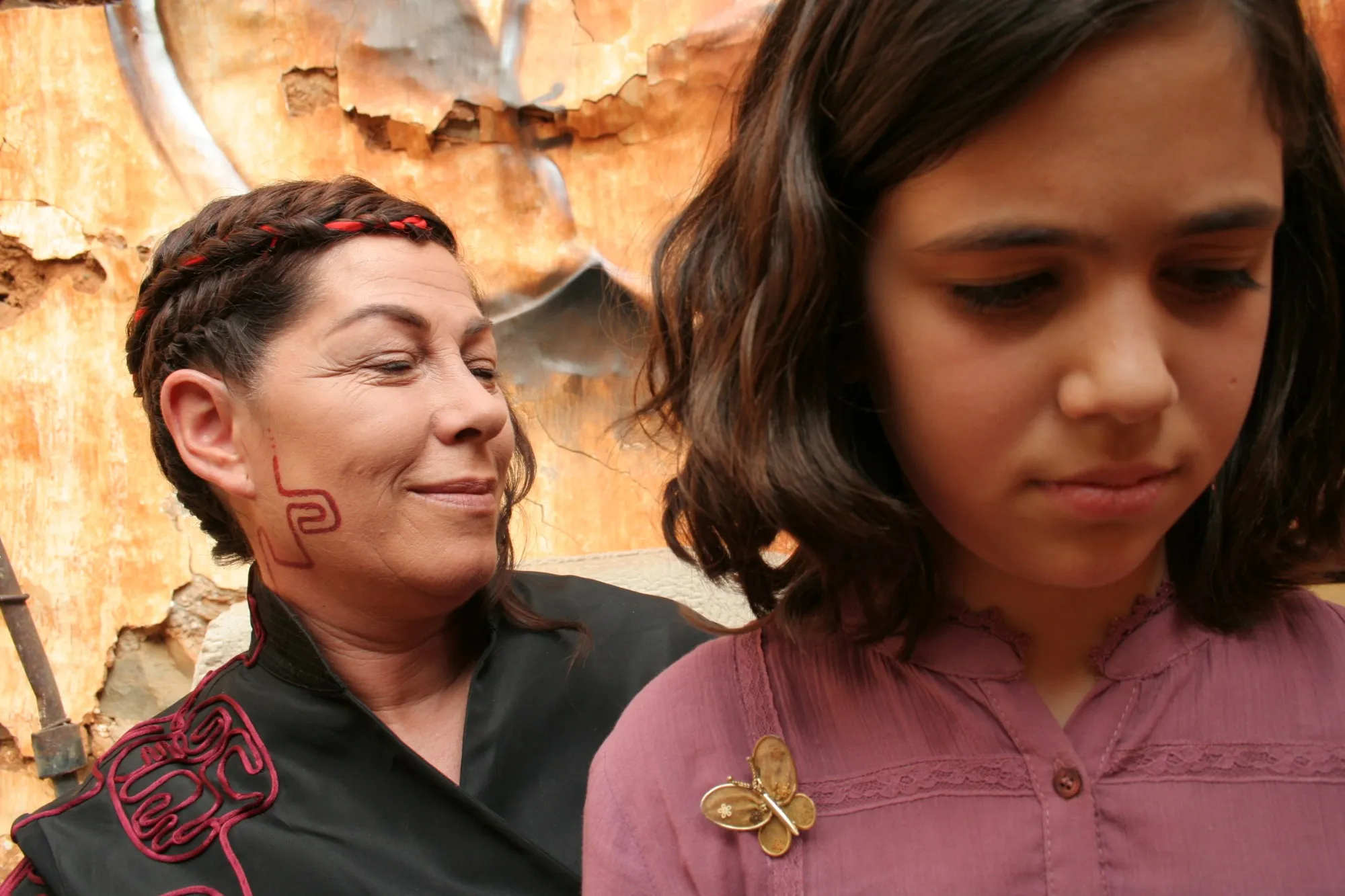 Mujer con trenzas y un dibujo rojo en la mejilla sonriendo detrás de una niña pensativa, ante una pared desconchada