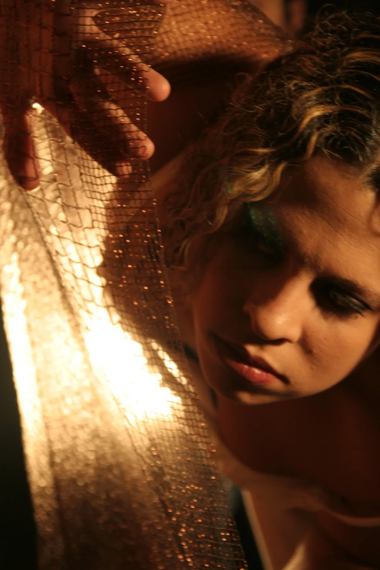 Mujer de pelo rizado con la mirada baja tras una malla dorada iluminada a contraluz