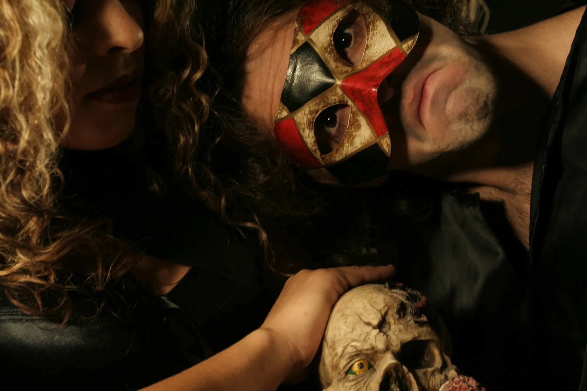 Pareja en penumbra, él con máscara de arlequín, junto a una calavera