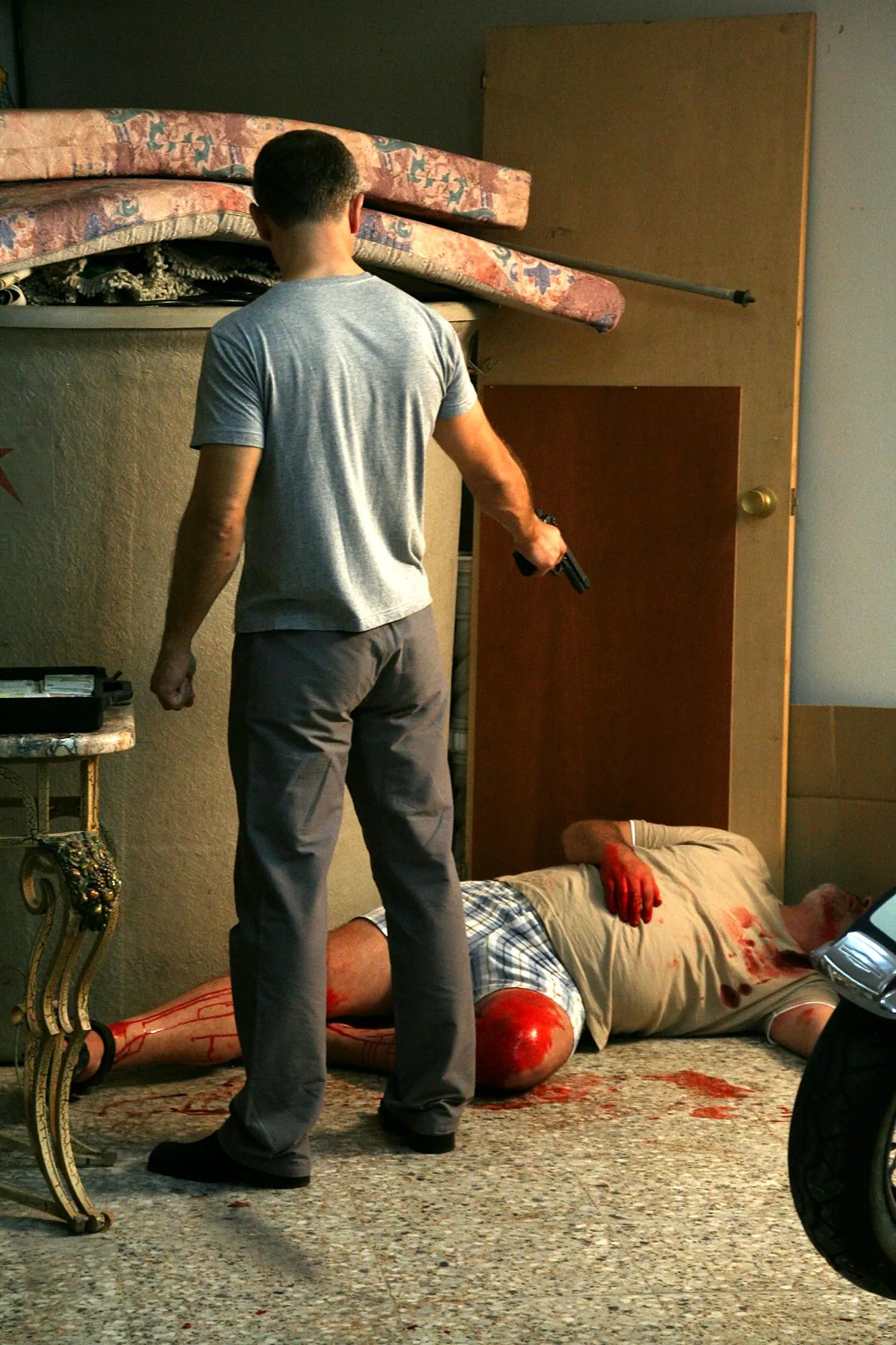 El actor de camiseta gris de pie con la pistola, junto a su víctima tendida en el suelo con sangre de atrezo