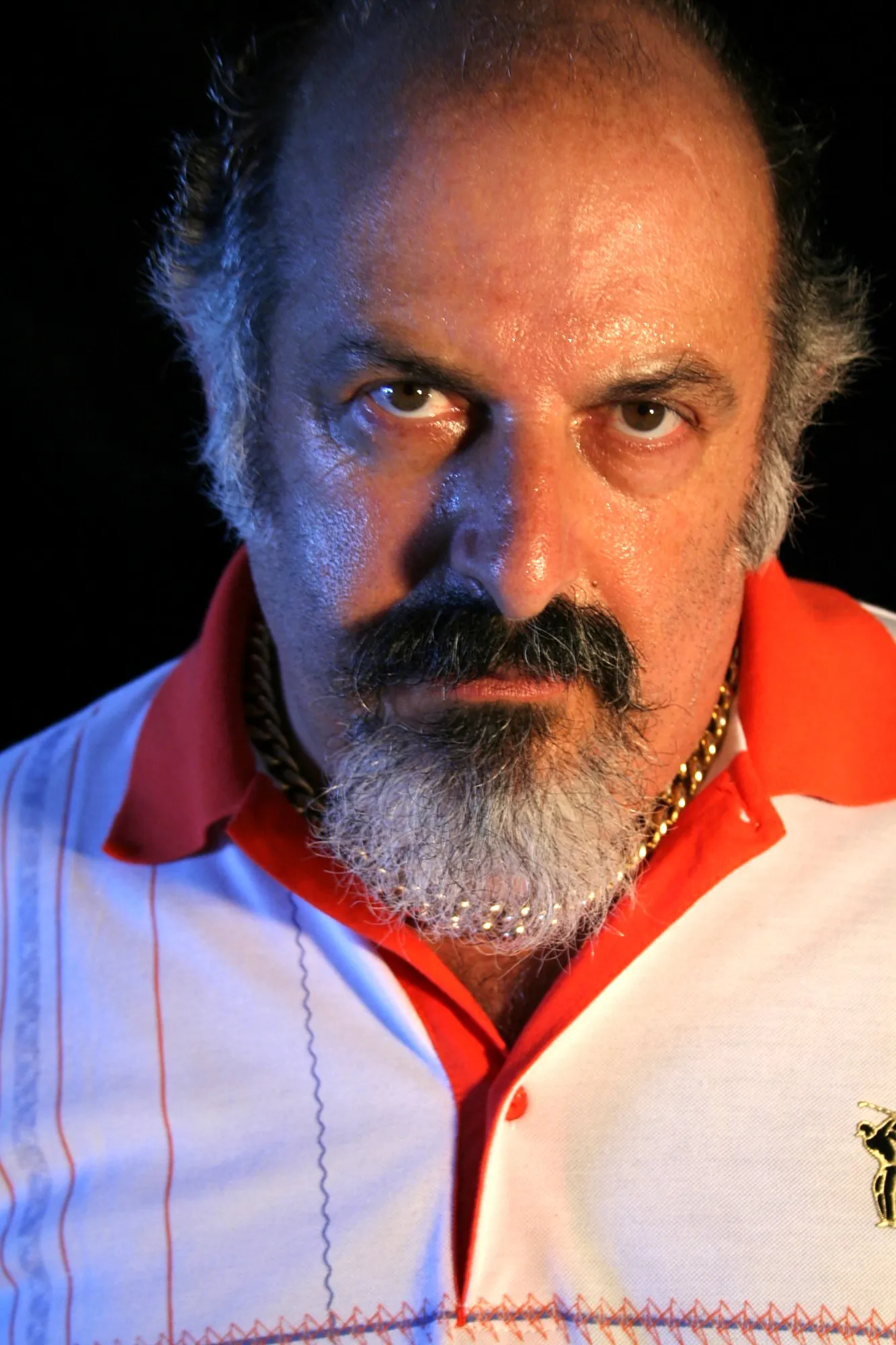 Actor de Granit con perilla, polo blanco y rojo y cadena dorada, mirando fijamente a cámara