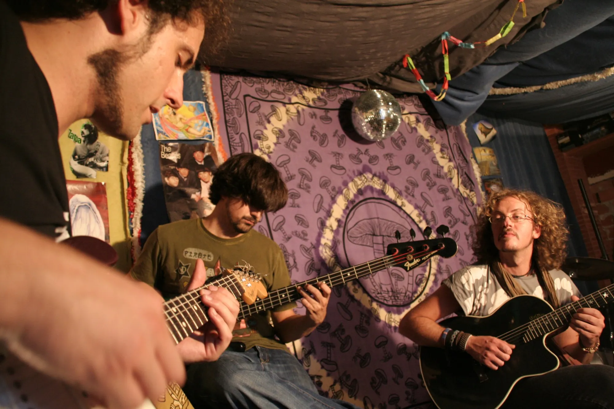 Tres músicos tocando guitarras y bajo en un local de ensayo con tapices y bola de espejos