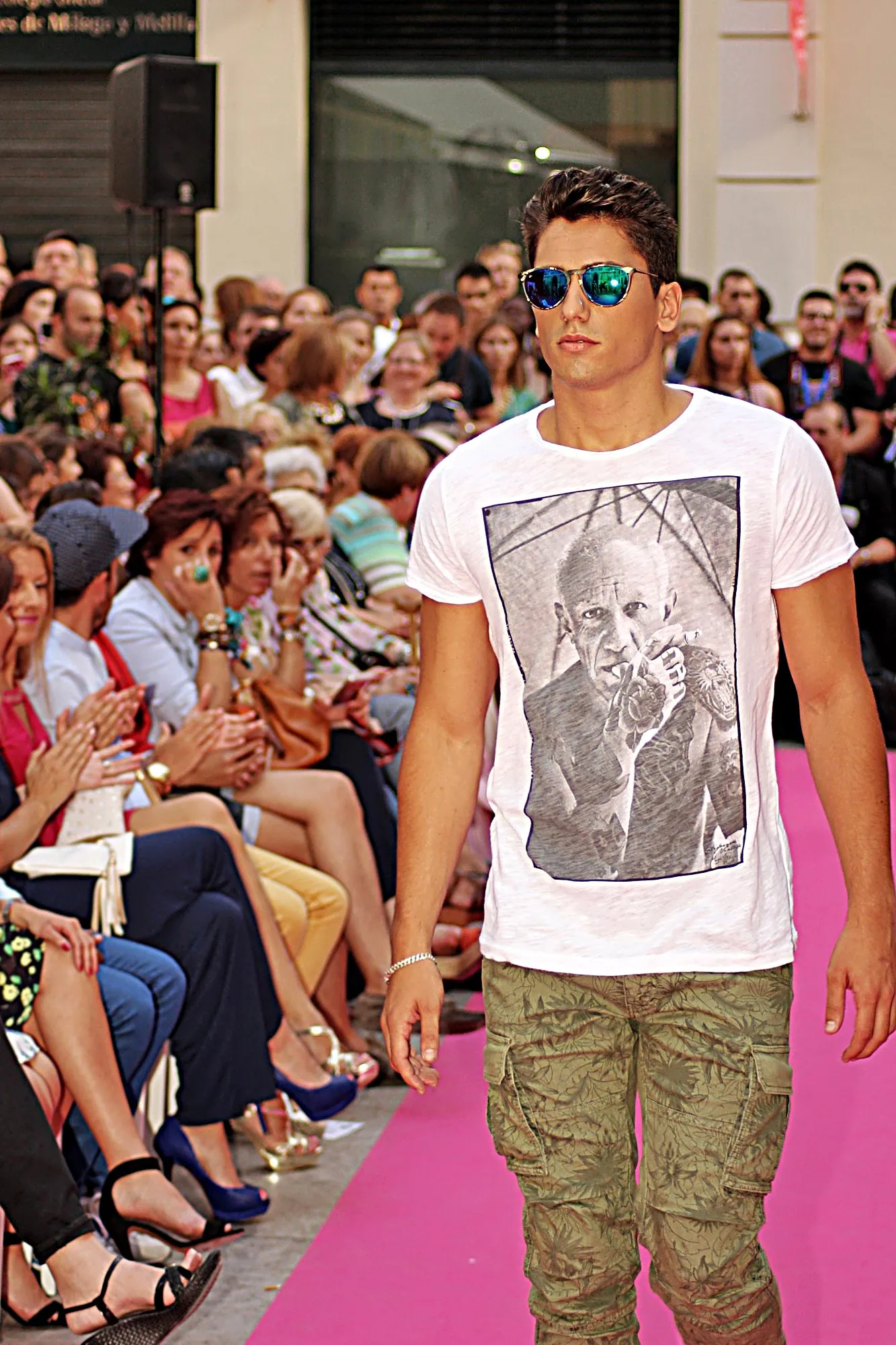 Modelo con gafas de espejo y camiseta blanca con un retrato en blanco y negro, junto al público sentado