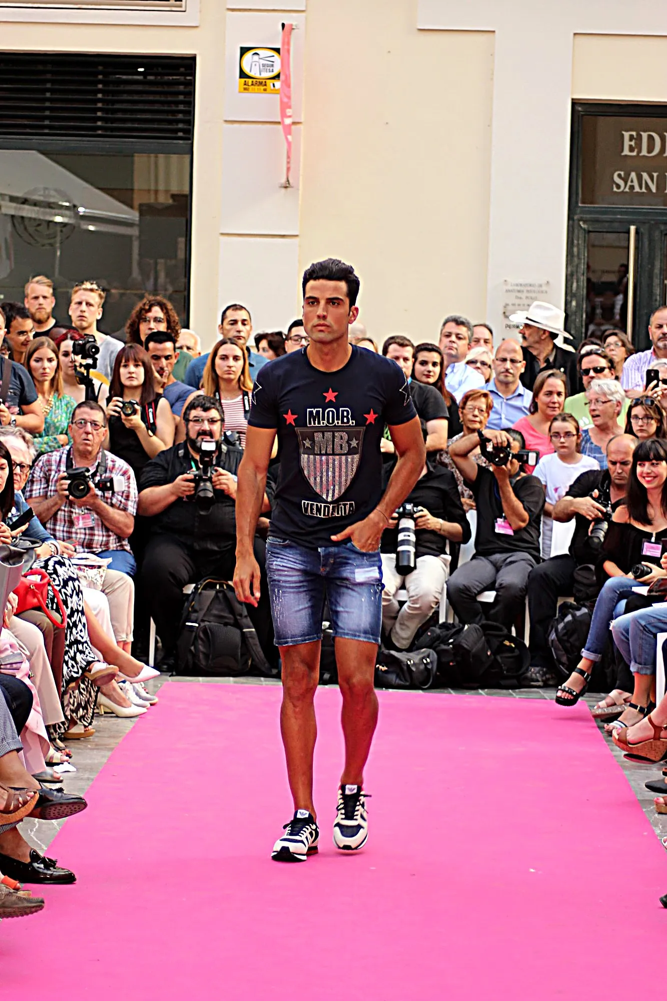 Modelo con camiseta azul marino de escudo y estrellas rojas y bermudas vaqueras en la alfombra rosa