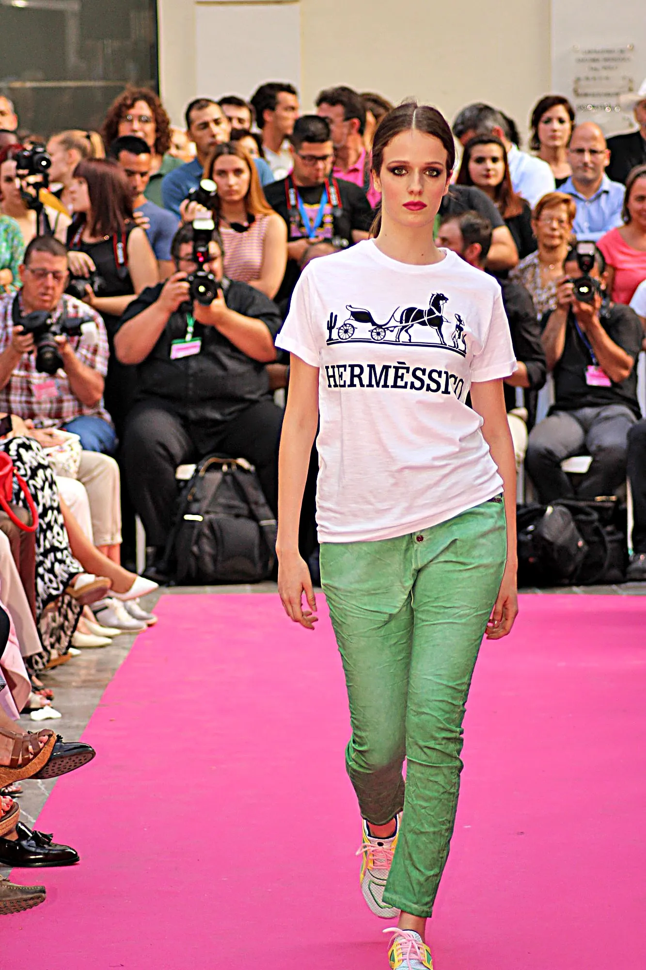 Modelo con camiseta blanca estampada y pantalón verde avanzando por la alfombra rosa