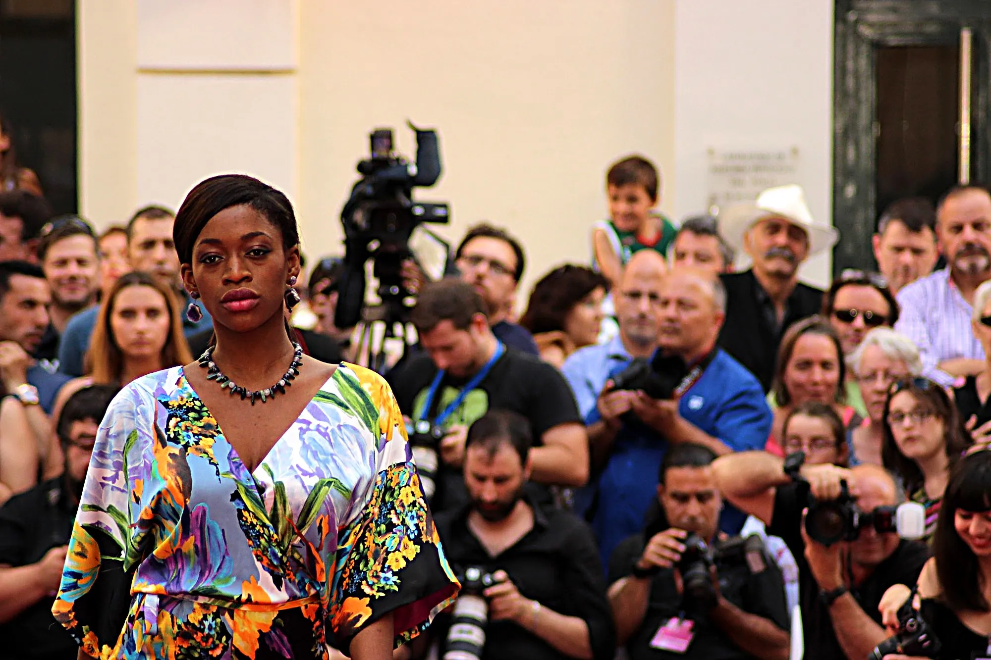 Modelo con kimono de flores y collar de piedras, con el público y una cámara de televisión detrás