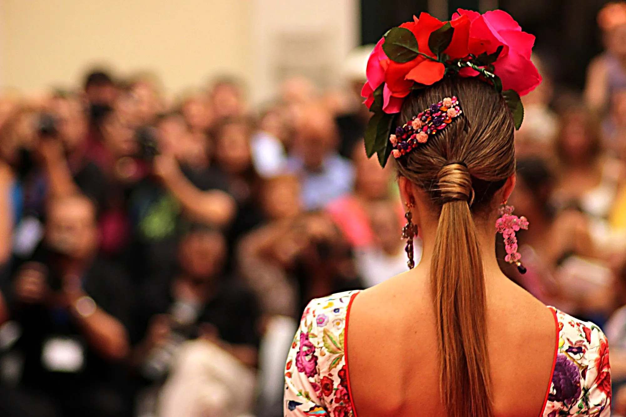 Espalda de una modelo con coleta, flores rojas y fucsias y una peineta de pedrería, frente al público desenfocado