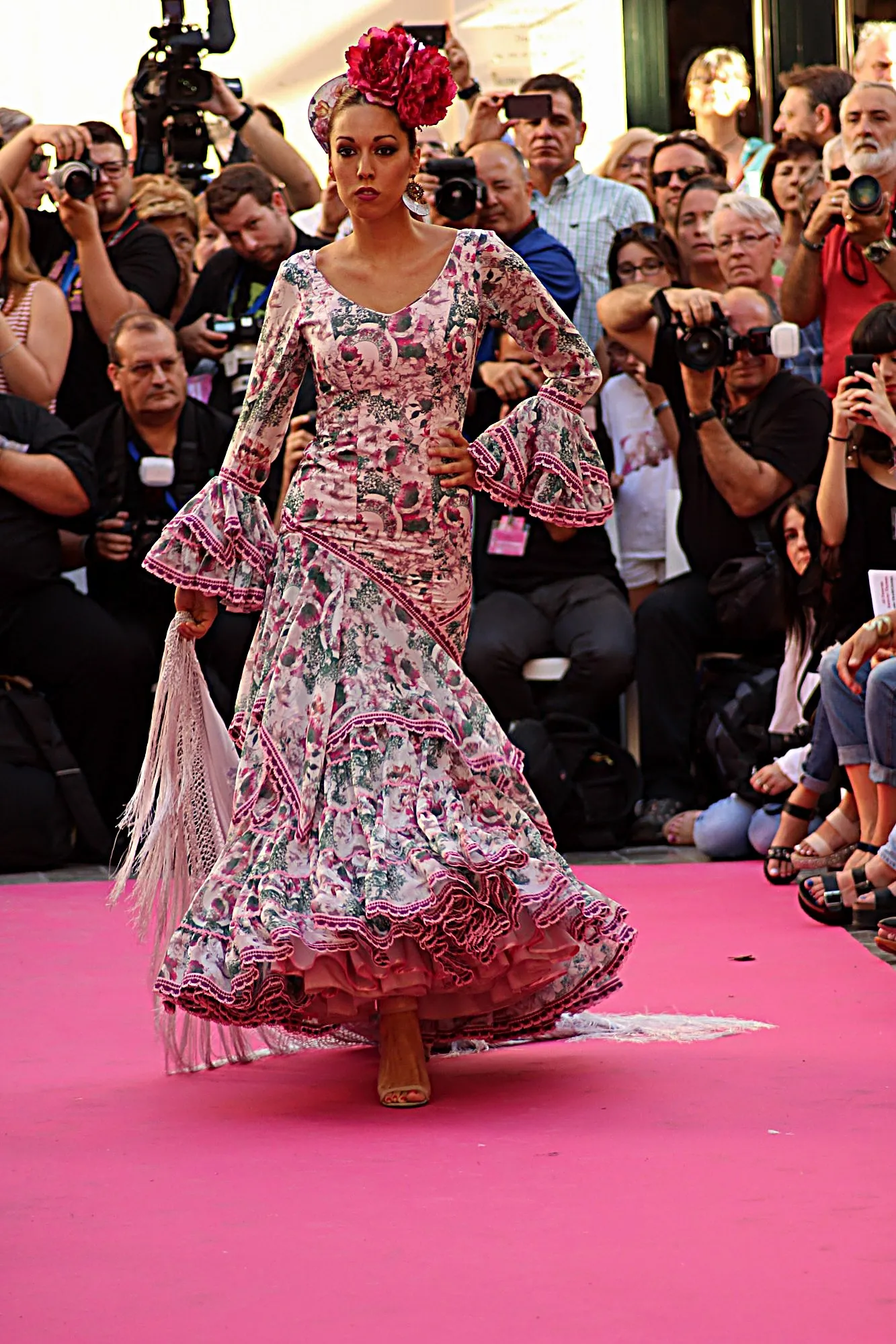 Modelo con traje de flamenca rosa de flores y mantón en la mano, avanzando por la alfombra rosa