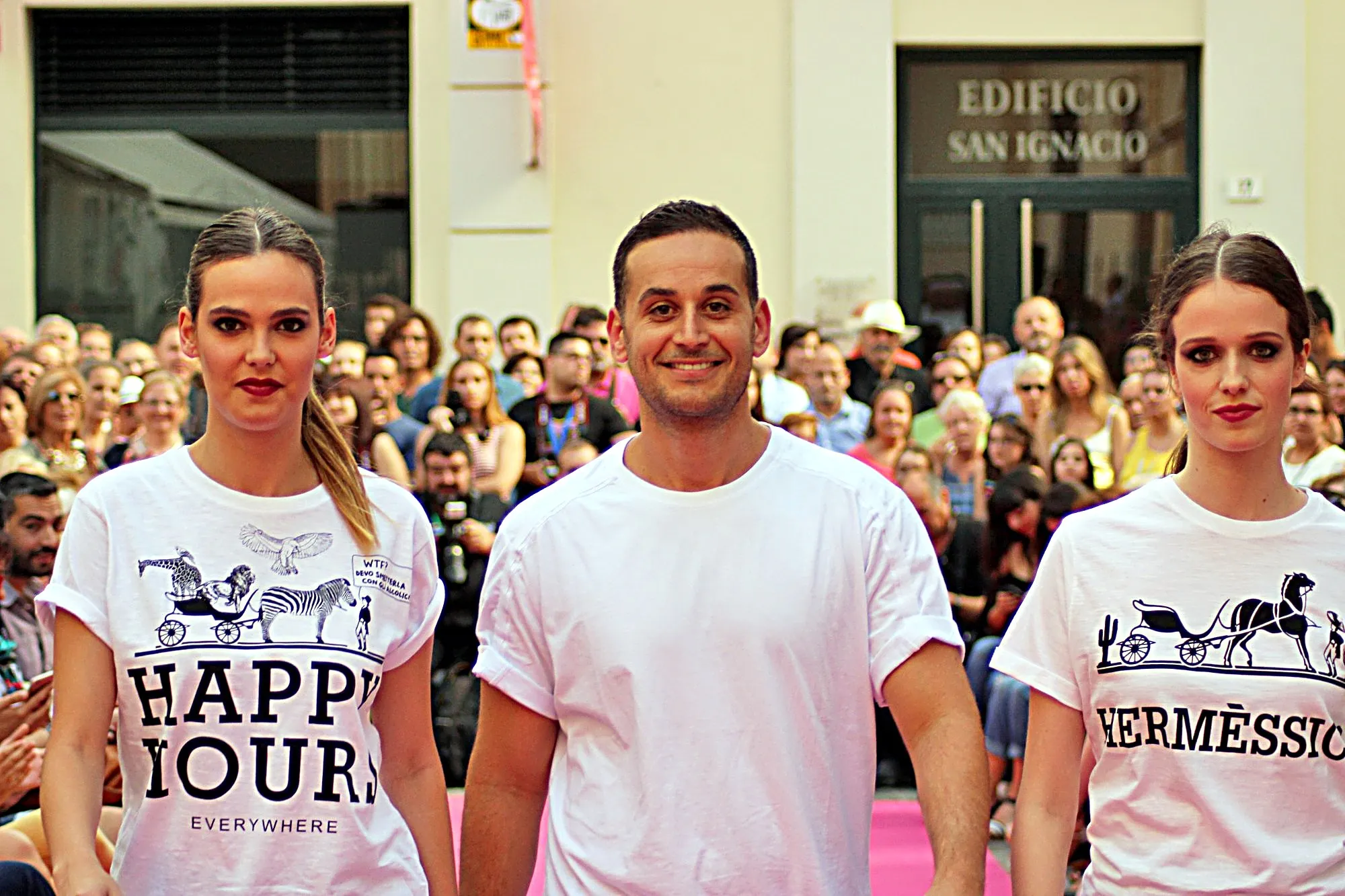 Diseñador sonriendo entre dos modelos con camisetas blancas estampadas, en el saludo final