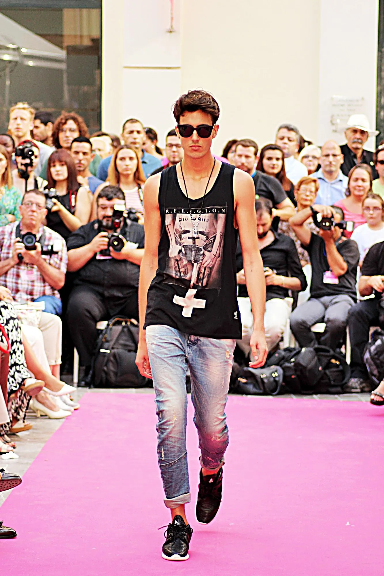 Modelo con camiseta de tirantes negra estampada, vaqueros y gafas de sol, avanzando por la alfombra rosa