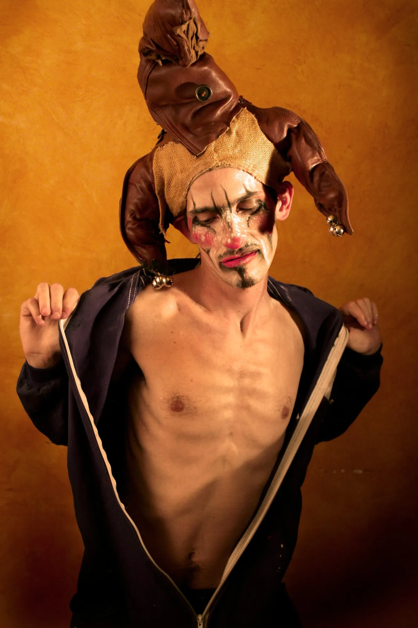 Actor con gorro de bufón y maquillaje triste abriéndose la chaqueta sobre fondo ocre
