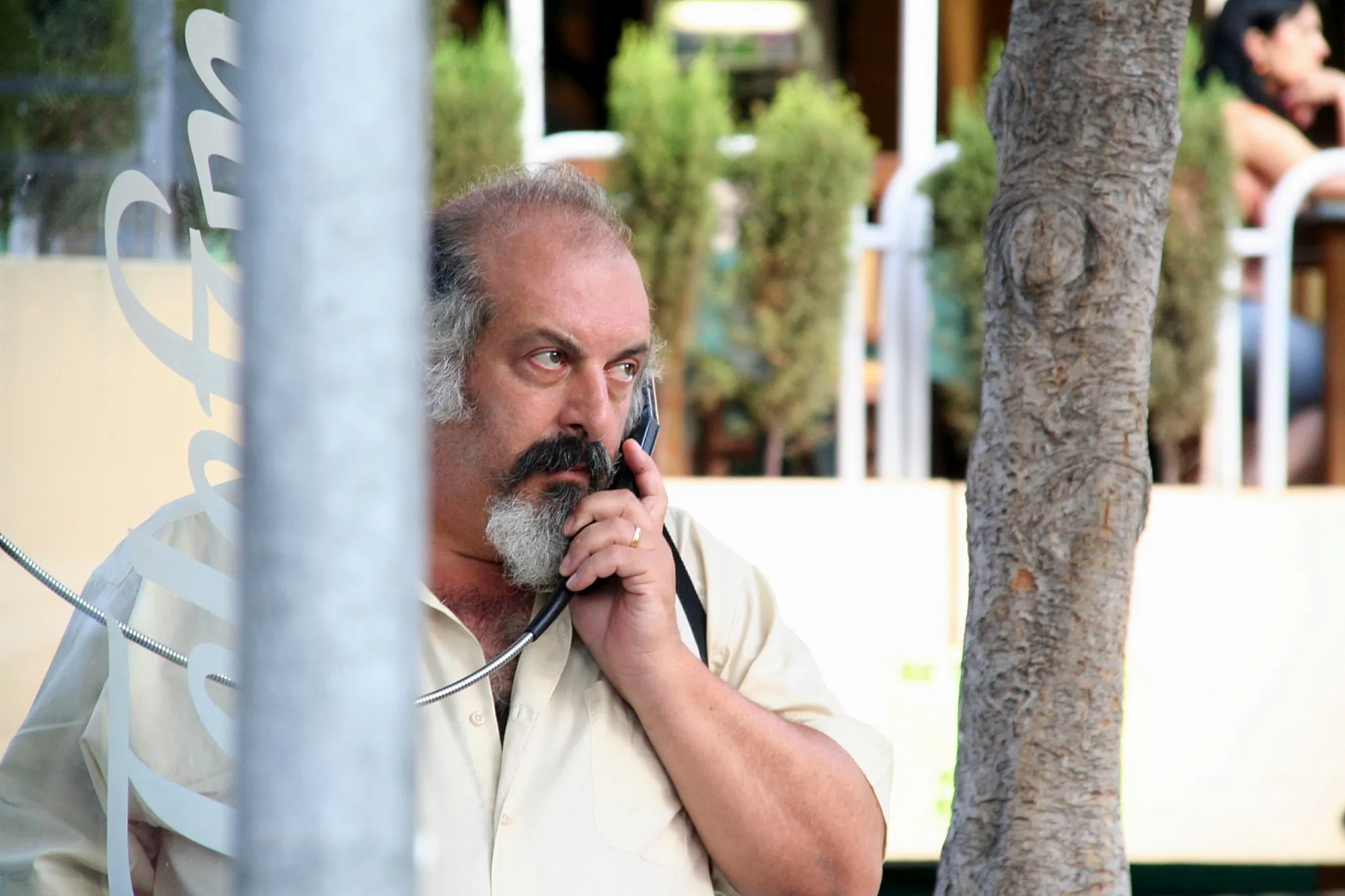 Actor con barba hablando desde una cabina de teléfono, entre un poste y un árbol