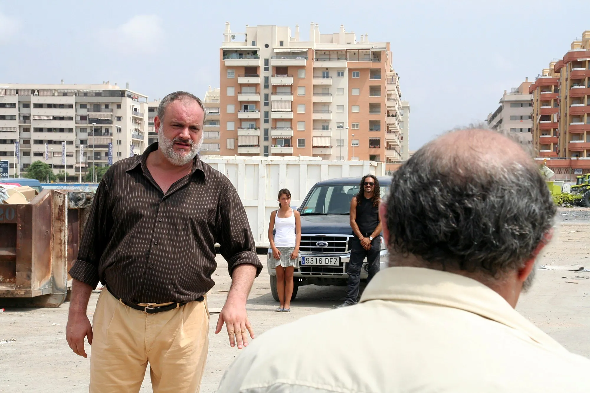 Escena en un descampado frente a los bloques de pisos, con dos actores al fondo junto a una camioneta