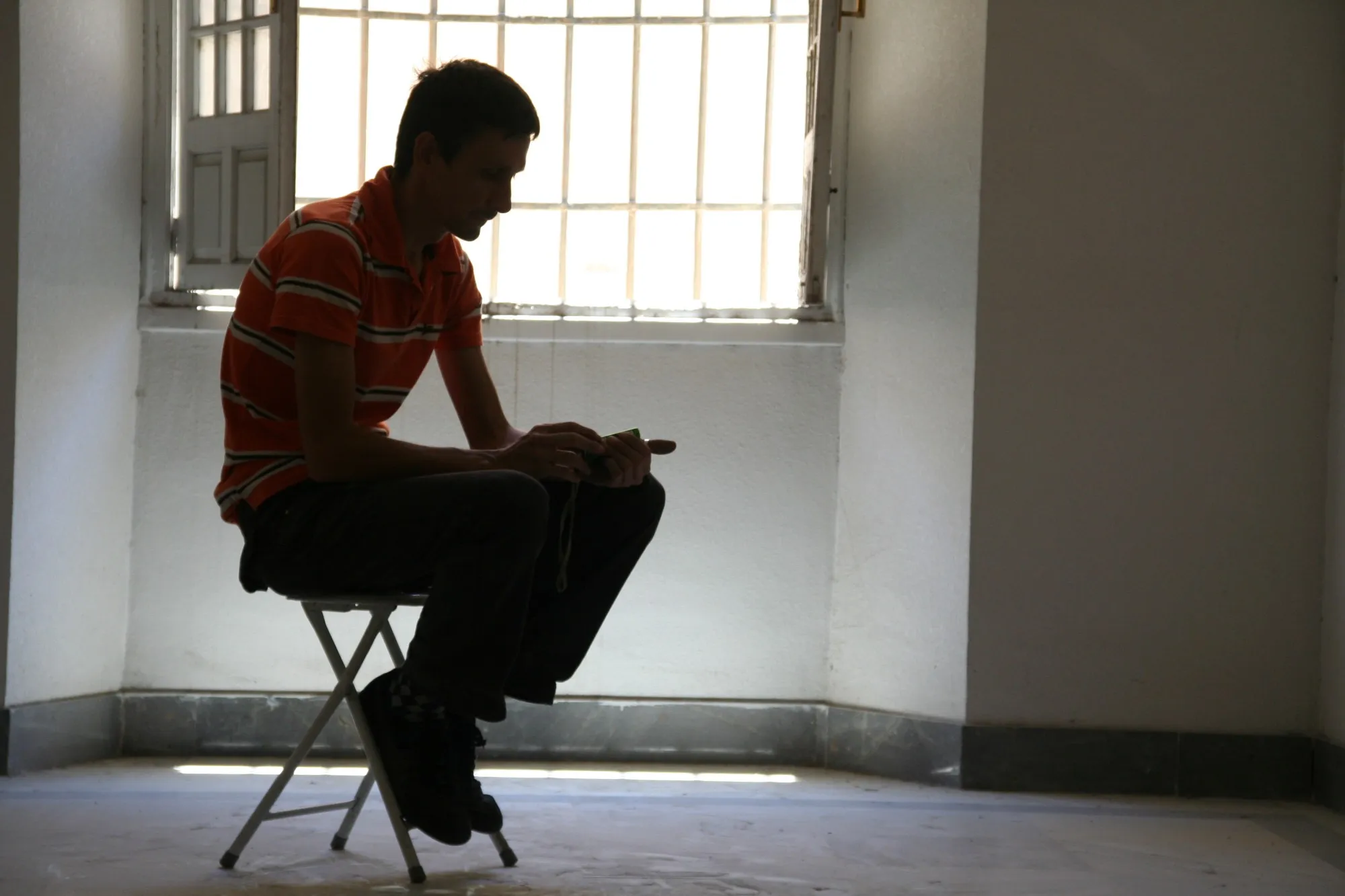 Hombre sentado en un taburete a contraluz frente a una ventana con rejas